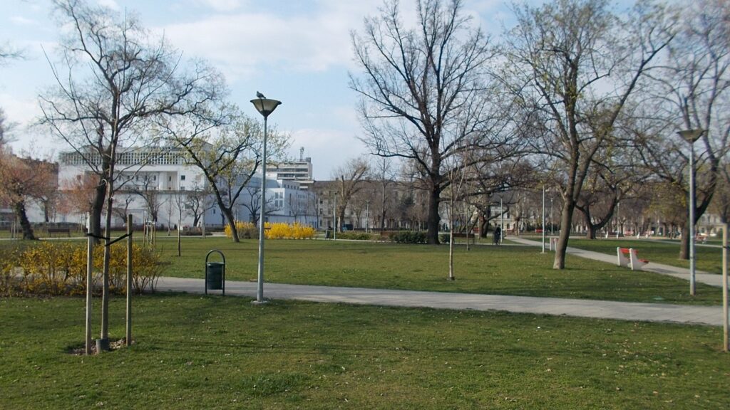 A II. János Pál pápa tér parkja Józsefvárosban, sétányok és fák, háttérben bérházakkal