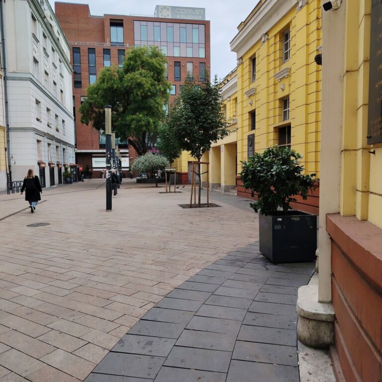 A Corvin köz napközben: járókelők a térkövezett gyalogos utcán, jobbra a Corvin mozi sárga épülete, háttérben a Corvin Plaza