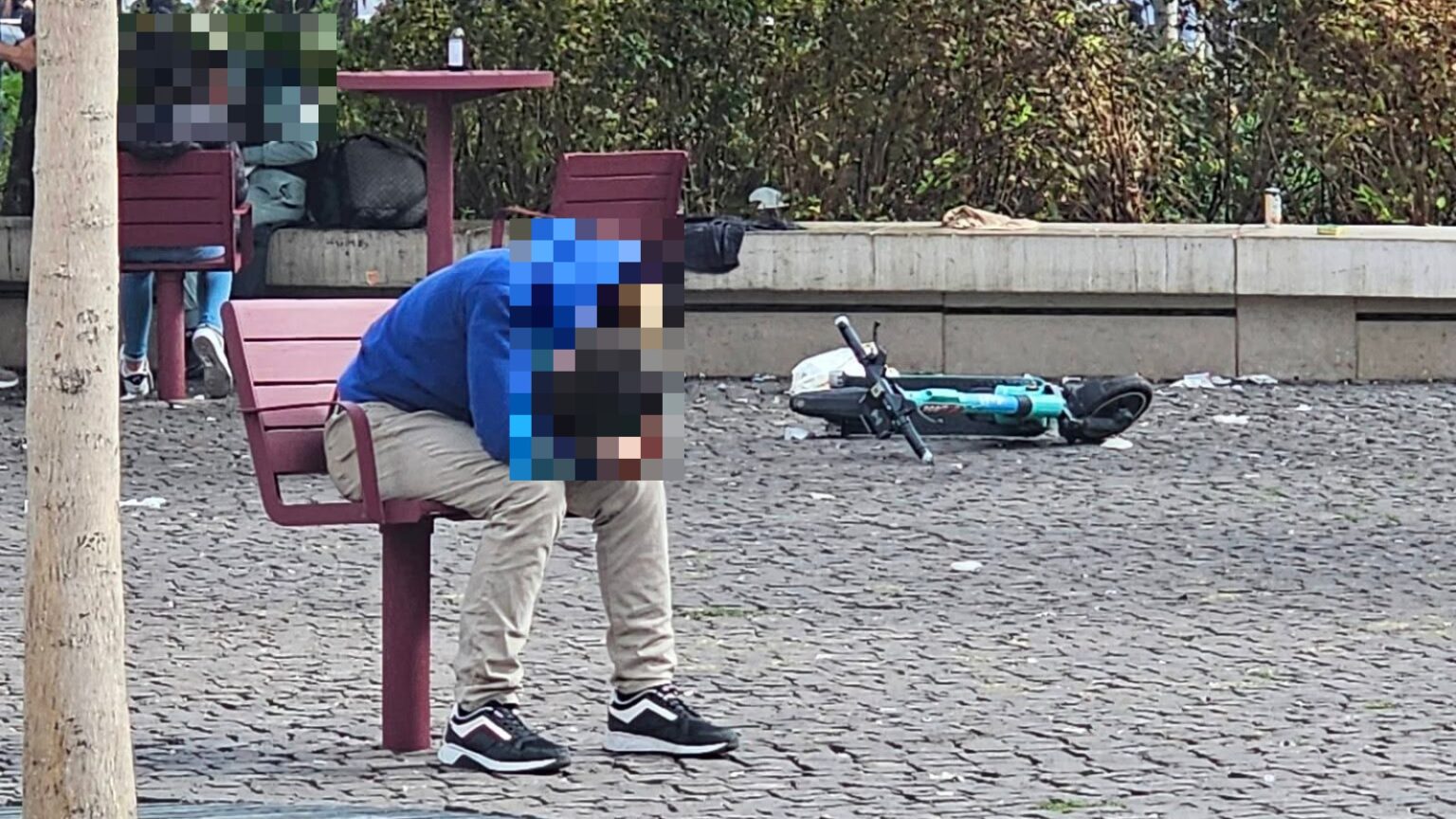 Kék pulóveres férfi ül előrehajolva a Blaha Lujza tér egyik székén, mögötte eldobott roller és szemét a kövezeten