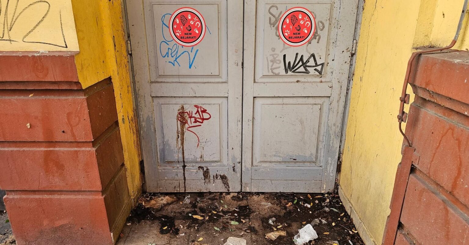 Graffitivel összefirkált, foltos szürke ajtó a Corvin közben, előtte szeméttel és sötét lerakódással borított küszöb